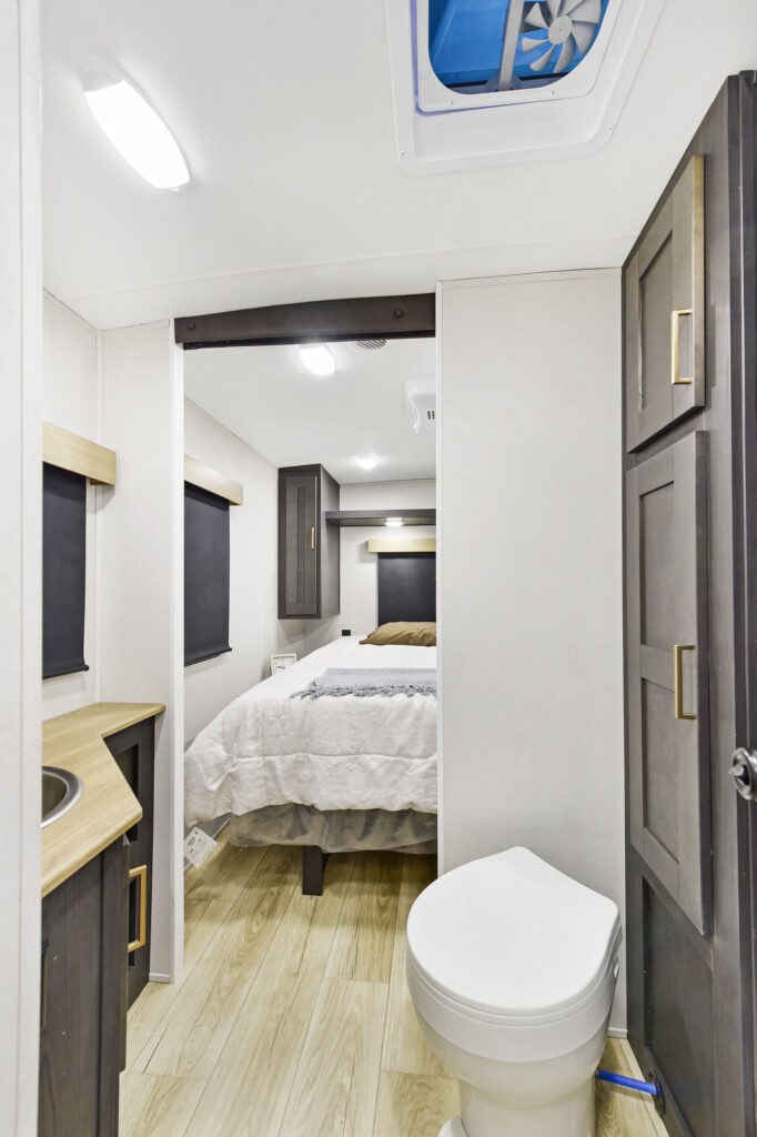 A compact RV interior with a white toilet in the foreground, wood-tone cabinets, a small sink, and a bed with white bedding in the background. Light wood flooring and overhead lighting are visible.
