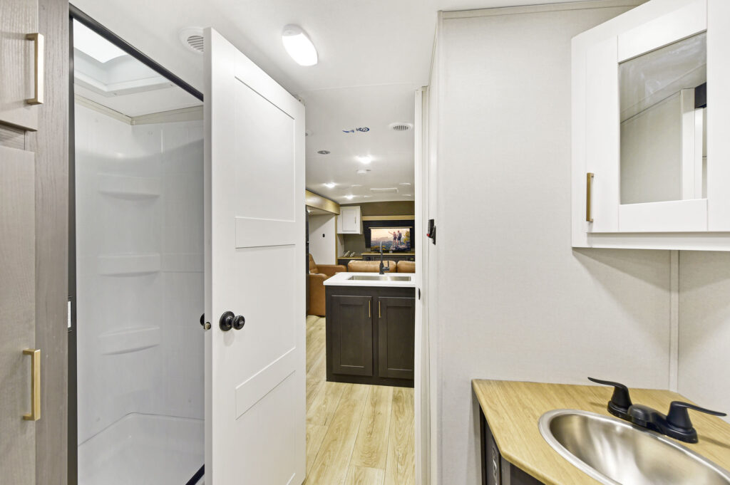 Modern RV interior showing a compact bathroom with a shower, white door, small vanity with a sink, and a view into the main living area featuring a kitchen and seating.