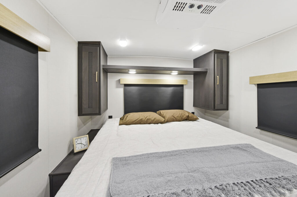 A modern RV bedroom with a neatly made bed, two brown pillows, a gray blanket, built-in cabinets, black-out shades on windows, and a small clock on the nightstand under bright ceiling lights.