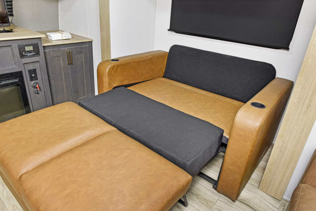 A brown leather sofa bed with black cushions is partially unfolded in a modern room with wood flooring, gray cabinets, and a black window shade.