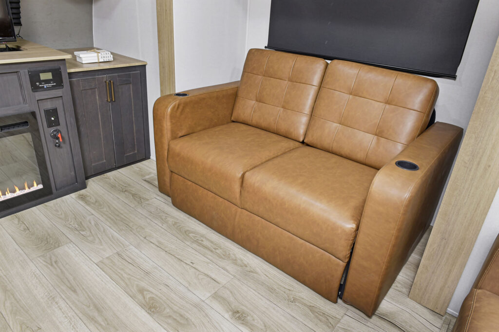 A brown leather loveseat with cup holders on each armrest sits on light wood flooring next to a dark wood cabinet and an electric fireplace in a modern living space.