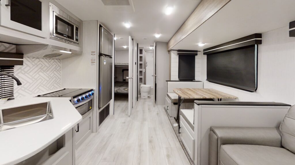 Interior of a modern RV featuring a kitchenette with a stove, oven, microwave, and sink on the left. A dining table with cushioned benches, a sofa, and a view towards a bedroom and bathroom are on the right. Light wood flooring and contemporary design.