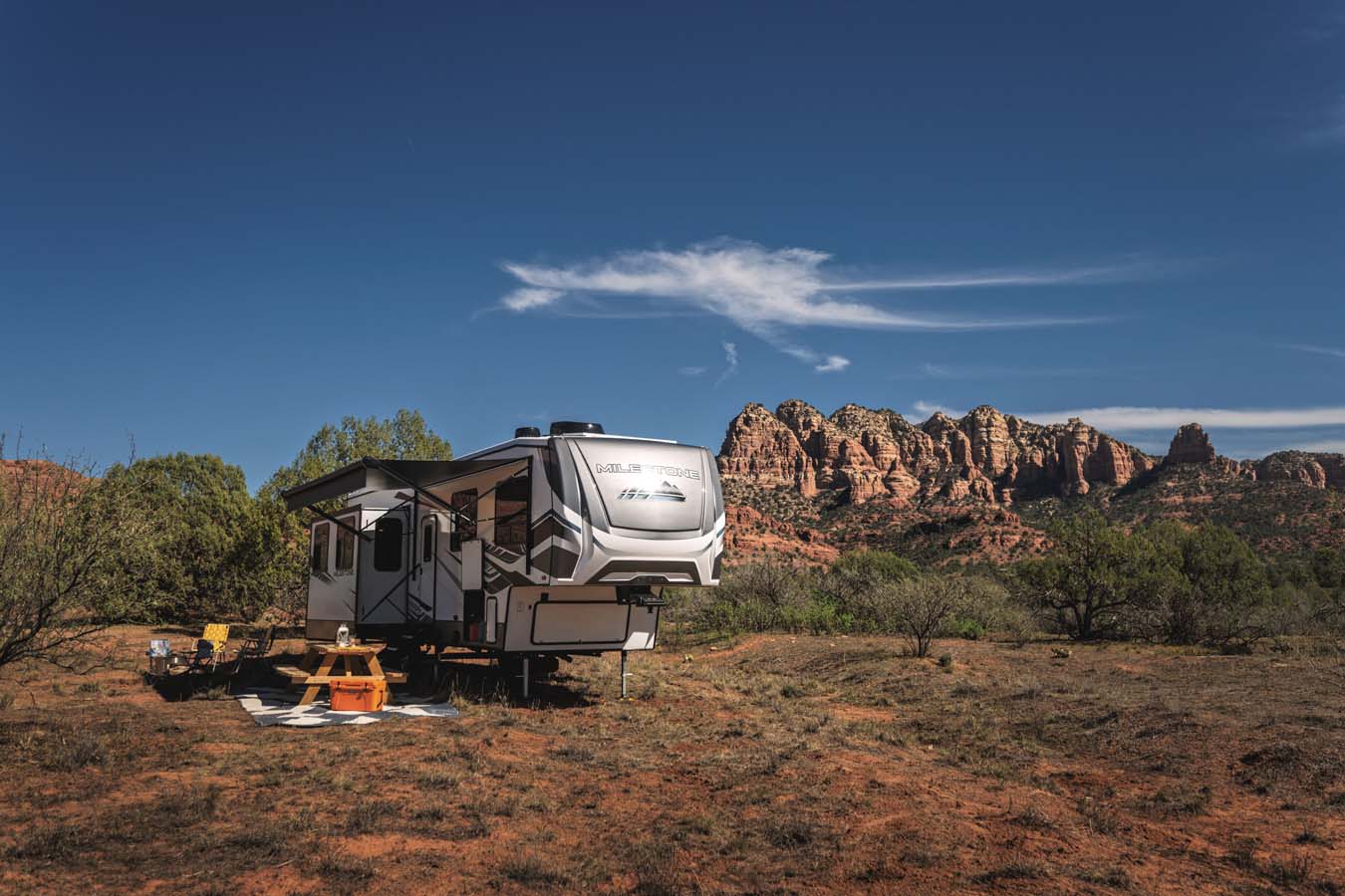 A RV is parked on a dirt patch in a desert landscape with red rock formations in the background. There's a picnic table with chairs and a rug nearby, surrounded by sparse vegetation under a clear blue sky.
