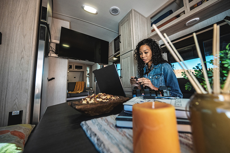 A woman sits at a table inside an RV, working on a laptop. The space is well-lit, with a TV mounted on the wall and a decorative candle in the foreground. Shelves and a window with a view complete the cozy interior.