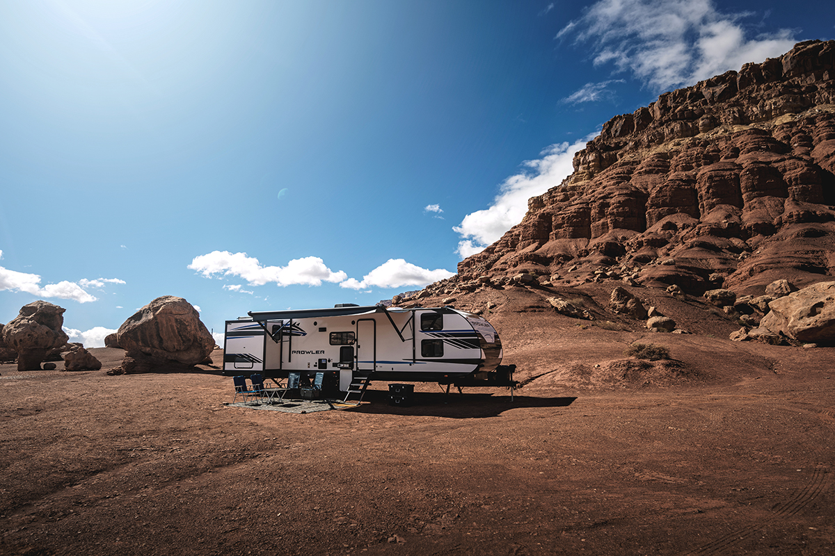 A travel trailer is parked on a barren, rocky landscape under a bright blue sky with a few clouds. Rugged cliffs rise on the right, and large boulders are scattered around, creating a dramatic desert scene.