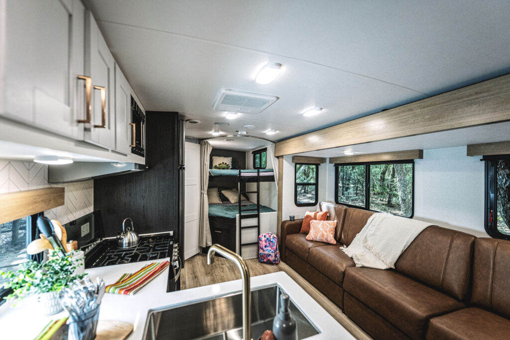 Interior of a modern RV featuring a cozy seating area with a brown leather sofa, a kitchen with white cabinets and a sink, a stove, and bunk beds in the background. Large windows add natural light and views of trees outside.