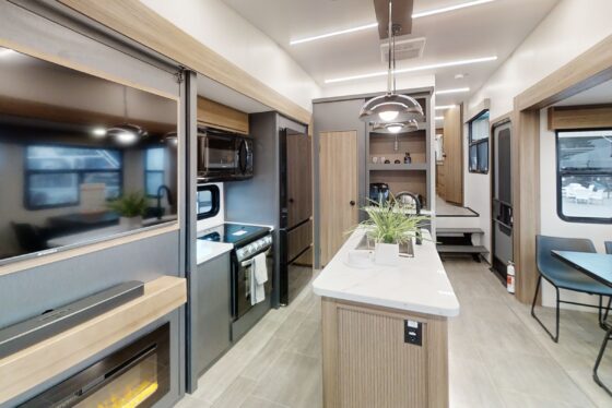 Interior view of the 2024 Heartland Corterra fifth-wheel RV living room, featuring plush seating, a TV, and an open kitchen area with modern appliances.