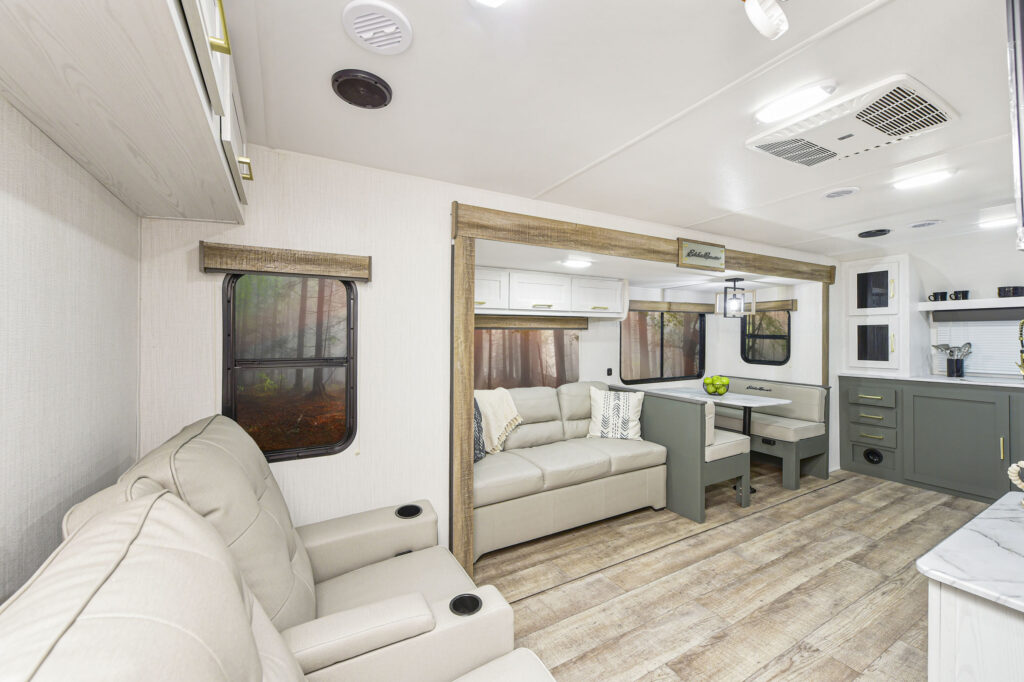 Modern RV interior with light wood floors, two beige recliners, a gray sofa, small dining table with benches, and kitchen area. Large windows show a forest view, and decor is light and minimalist.