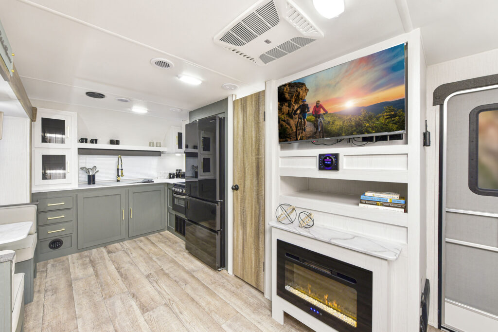 Modern RV interior with light wood flooring, green kitchen cabinets, black appliances, wall-mounted TV displaying a scenic image, electric fireplace below, and a door to the right. The space looks spacious and well-lit.