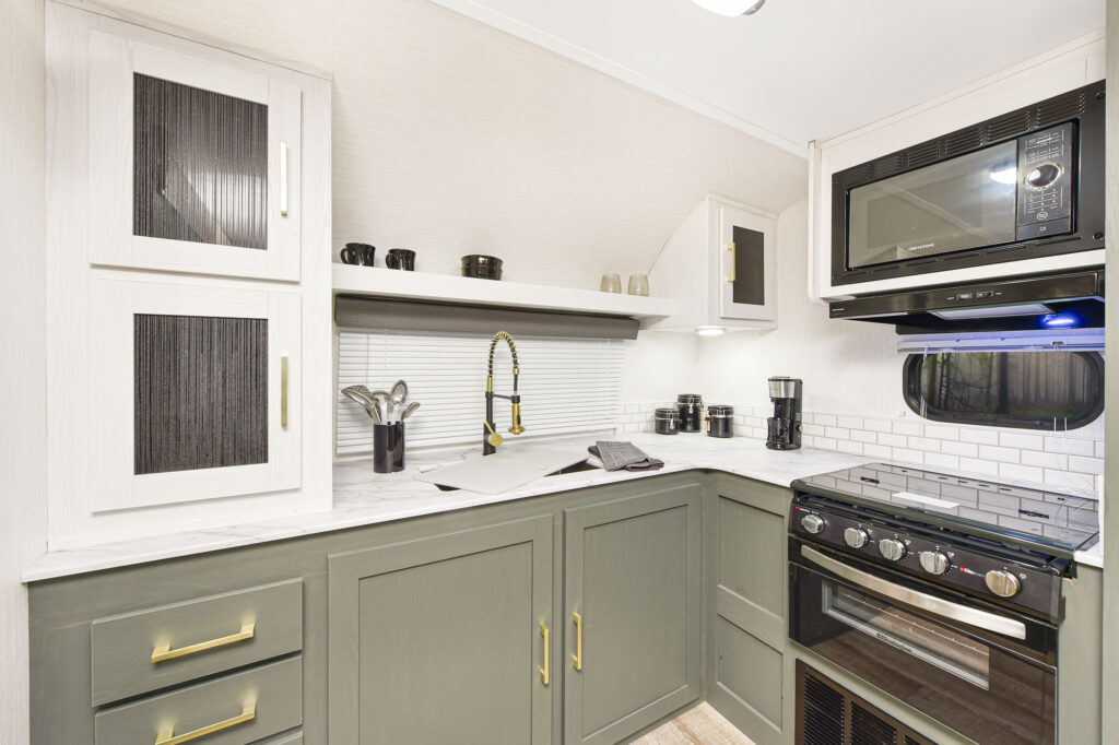 Modern RV kitchen with green lower cabinets, white upper cabinets, a black stove with oven, microwave, sink with gold faucet, kitchen utensils, and various cups and containers on the white countertop and shelves.