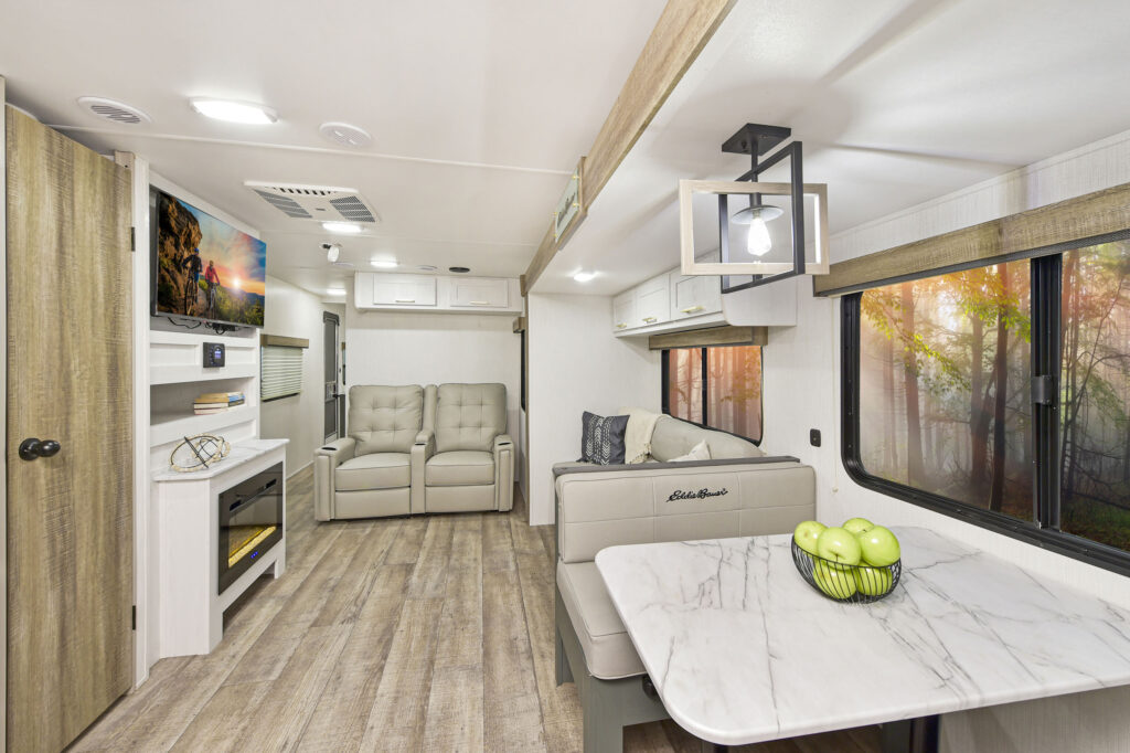 Modern RV interior featuring light wood floors, a marble-look dining table with green apples, a cozy couch, reclining chairs, a wall-mounted TV above a fireplace, and large windows showing trees outside.