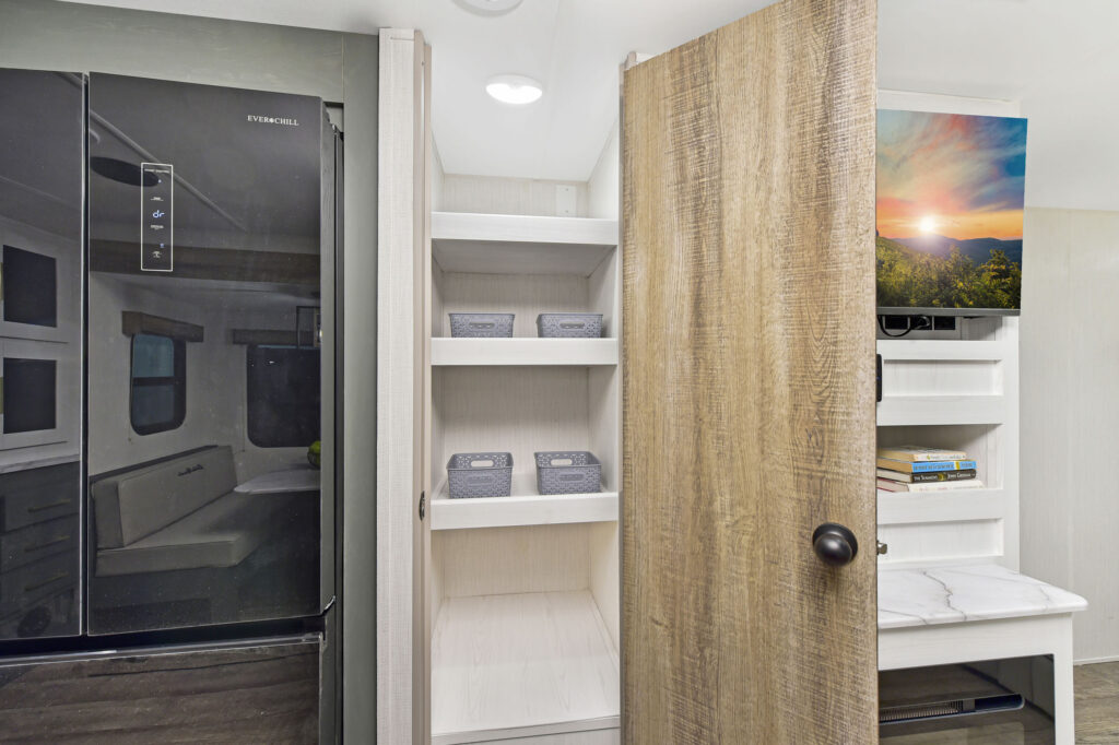 A modern RV interior featuring a black refrigerator on the left, white shelving with storage bins in the center, and a partially open wooden door revealing a TV and shelves with books on the right.