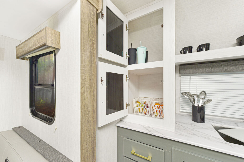 A kitchen area with open white cabinets showing mugs, cups, and a basket of fruit. Below is a marble countertop with utensils in a holder, near a window with blinds and another window with a valance.
