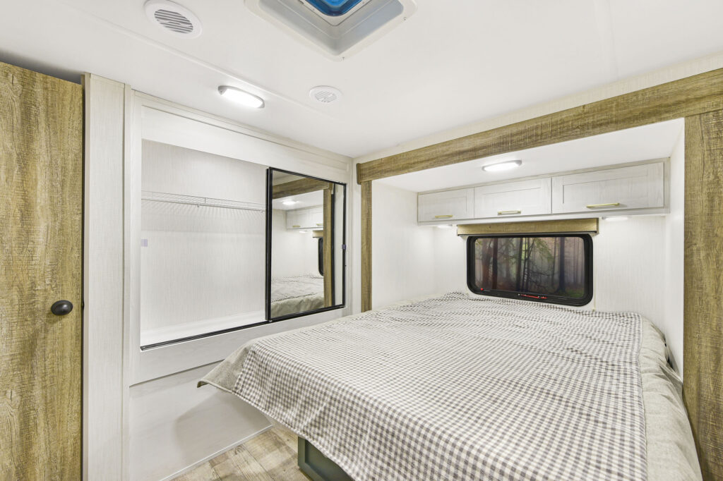 A modern RV bedroom featuring a checkered bedspread, built-in overhead cabinets, a window with a shade, wood accents, and a large mirrored closet door reflecting the interior.