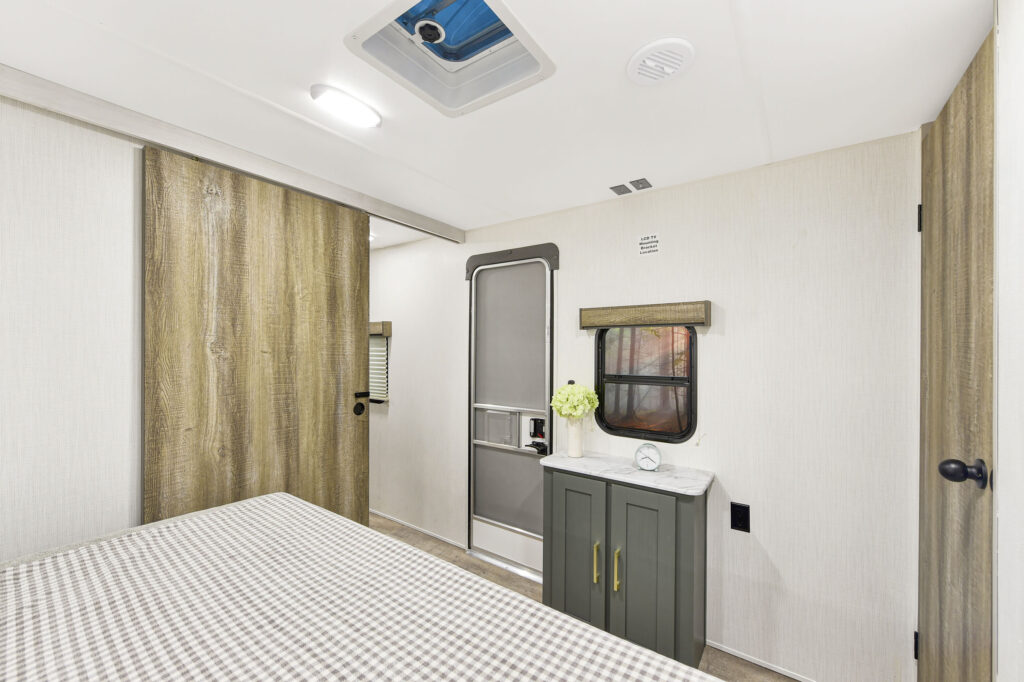 A small, modern RV bedroom with a checkered bedspread, wood sliding door, small green cabinet with flowers and clock, window with blinds, and a door leading outside. Ceiling light and vent are visible.