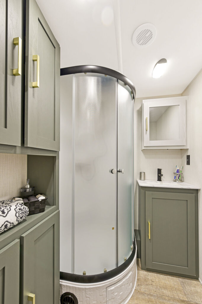 A modern RV bathroom features a curved frosted glass shower, green cabinets with gold handles, a sink with a green vanity, a mirrored medicine cabinet, and wood-look flooring.