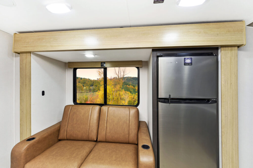 Interior of a modern RV featuring a brown leather sofa and a stainless steel refrigerator. Large window behind the sofa shows a scenic view of autumn trees. Natural wood accents and bright lighting create a cozy atmosphere.