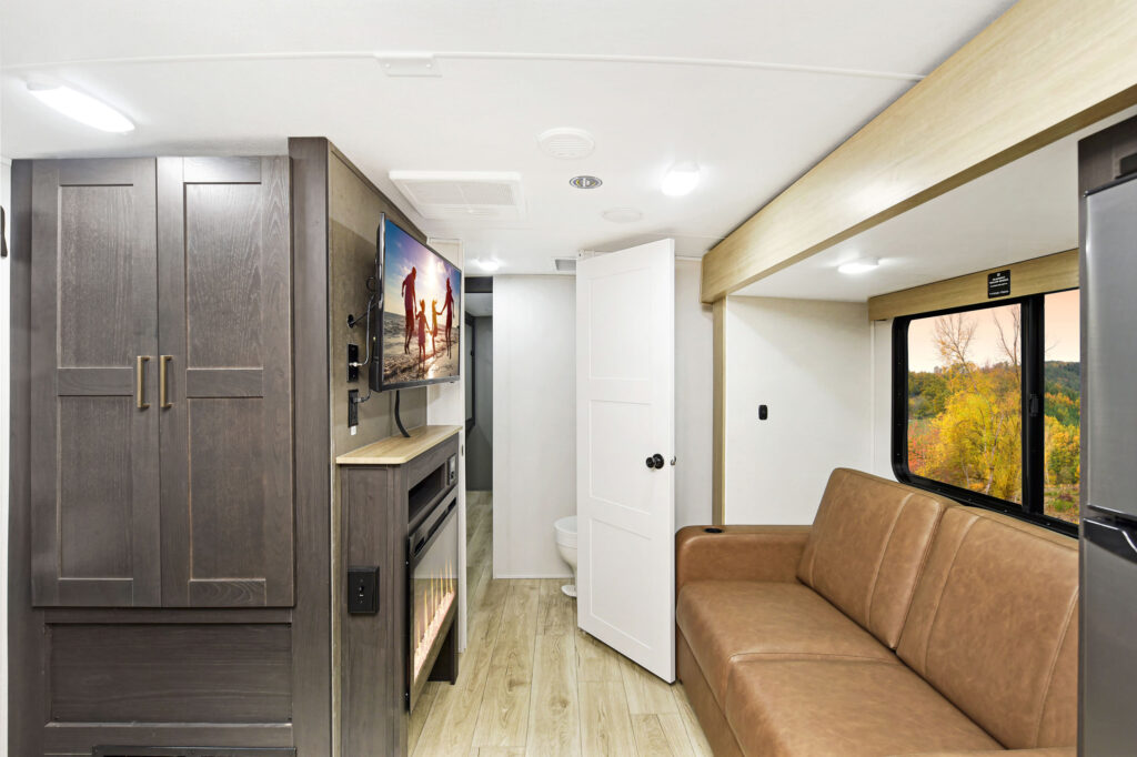 Interior of a modern RV with wooden flooring. On the left, there is a TV mounted above a fireplace, flanked by dark cabinets. A brown sofa and large window with a scenic view are on the right. A door at the back leads to a small bathroom.
