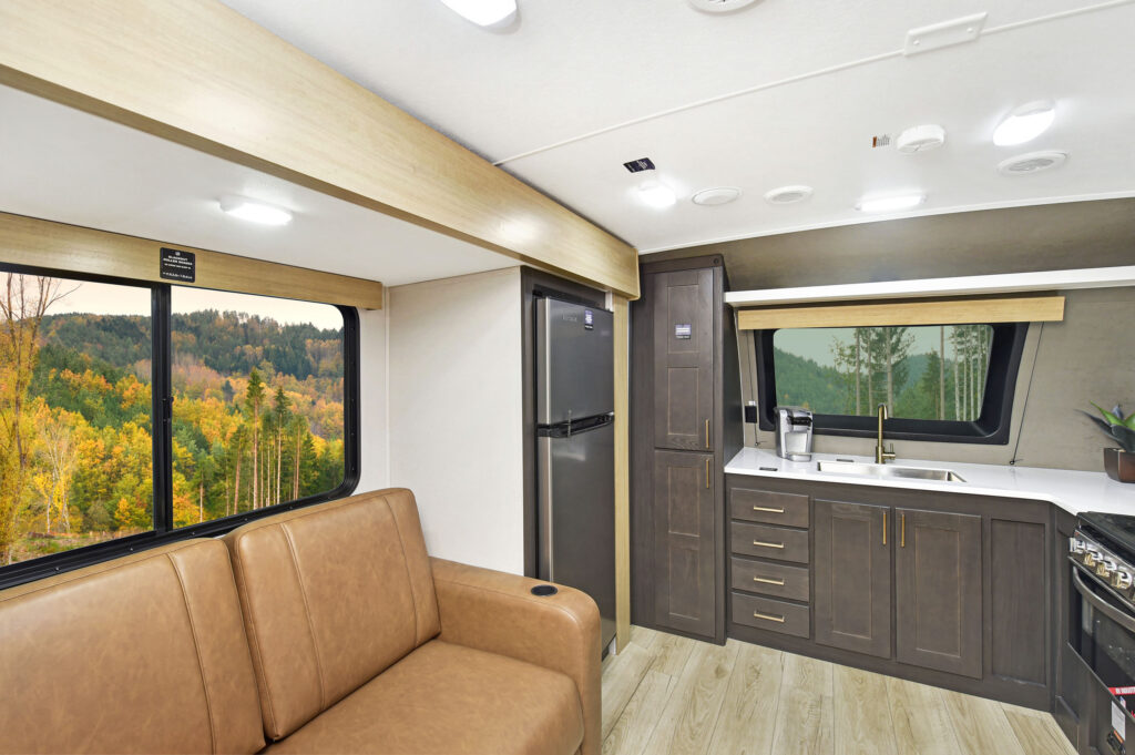 Interior of an RV featuring a brown couch, a kitchenette with a sink, stove, and refrigerator. Large windows reveal a forest with autumn foliage outside, creating a cozy and scenic atmosphere.