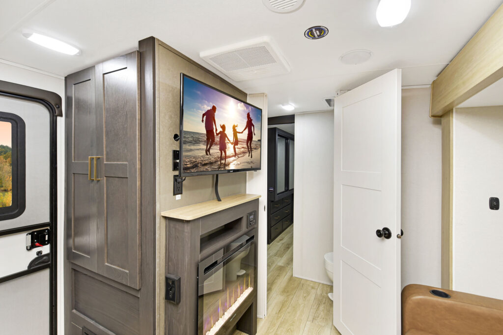 Interior of an RV featuring a modern fireplace, a mounted TV displaying a silhouette of a family at the beach, white walls, and wood accents. The space is bright with overhead lighting and minimalistic design.