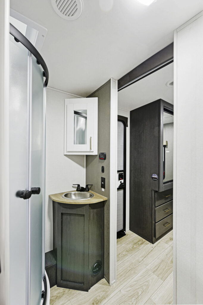 Interior of an RV bathroom featuring a shower with frosted glass doors on the left, a small sink with a wooden countertop and a mirrored cabinet above, and a dark wood door leading to another compartment. Light wood flooring is visible.