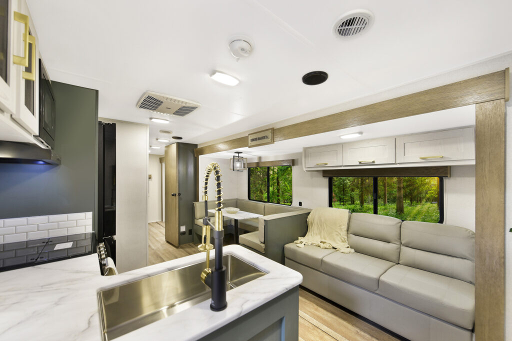 Modern RV interior featuring a marble countertop with sink, gray sofa, dining area, and large windows with forest views; neutral tones and overhead cabinetry create a spacious, cozy feel.