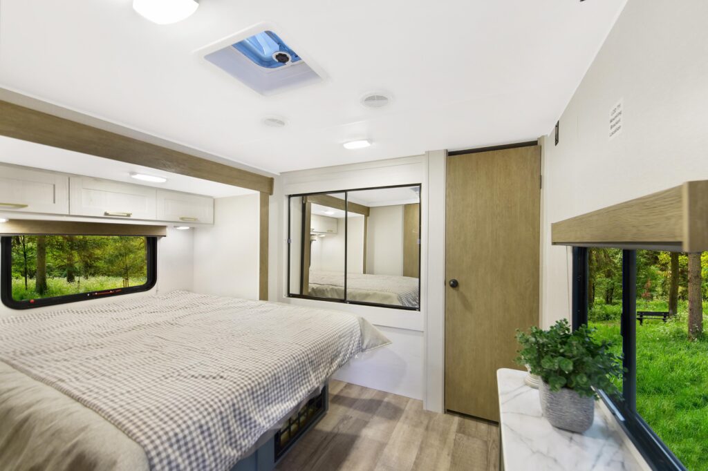Bright, modern RV bedroom with large windows showing a green forest, a neatly made bed with a beige comforter, wood accents, a mirrored closet, and a small plant on a marble-look shelf.
