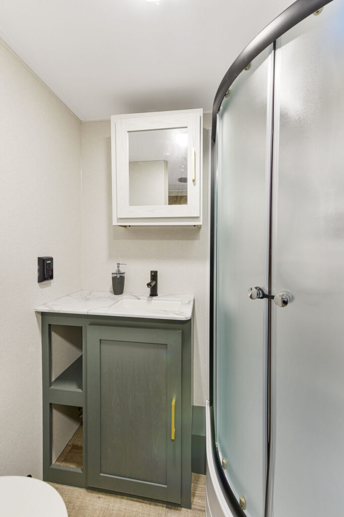 Small modern bathroom with a rounded frosted glass shower, gray vanity with a marble countertop, sink, mirror cabinet above, and beige walls.