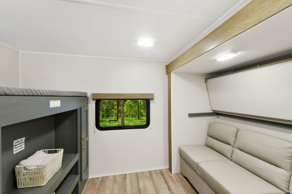 Modern RV interior featuring a gray sofa, a small window with a view of greenery, a bunk bed with a basket below, light wood flooring, and white walls with simple, bright lighting.