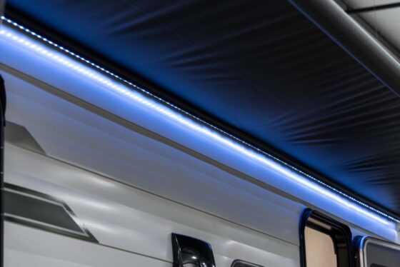 Close-up of an RV exterior with a black awning extended and a strip of blue LED lights illuminating the area beneath the awning. The RV has white and gray siding with geometric patterns.