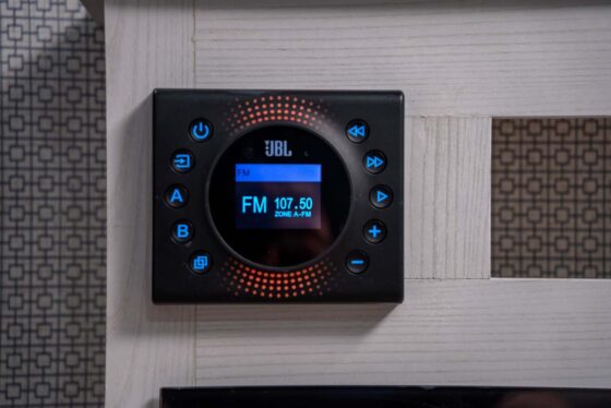 A black JBL wall-mounted audio control panel displays FM 107.50 ZONE 4-FM on its blue screen, with various buttons surrounding the circular display. The panel is installed on a light-colored wall.