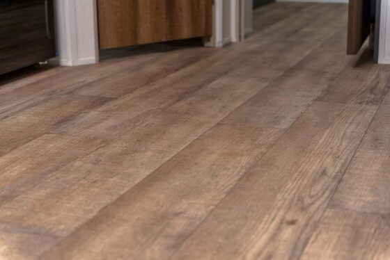 Close-up view of a hallway with wood-look vinyl plank flooring and doors partially open along the walls. The flooring features natural wood grain patterns in various brown shades.