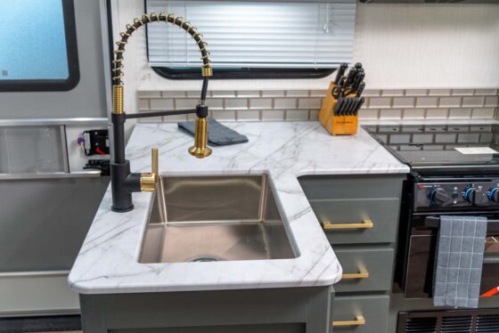 Modern RV kitchen with a marble countertop, stainless steel sink, gold and black faucet, knife block, stovetop, and drawers with gold handles. A towel hangs on the oven door, and a window is above the counter.