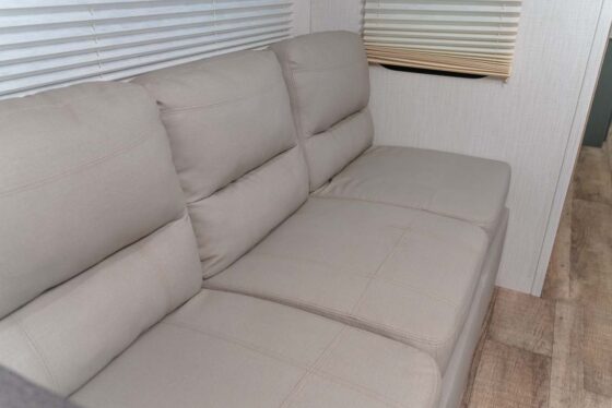 A beige, cushioned sofa with three seats sits against a wall with white blinds, on a light wood floor in a bright indoor space.