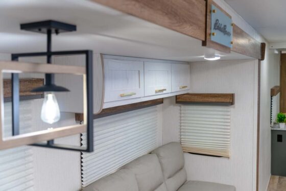 Modern RV interior featuring a beige sofa, white cabinetry with gold handles, wood accents, window blinds, and a decorative ceiling light fixture with a visible bulb.