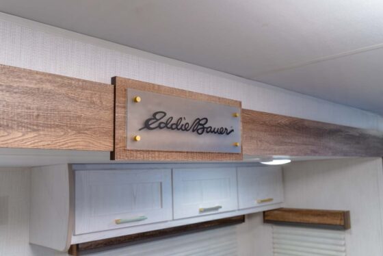A wooden beam inside a room features a sign with Eddie Bauer written in script, mounted with gold fasteners above white cabinets and a small ceiling light.