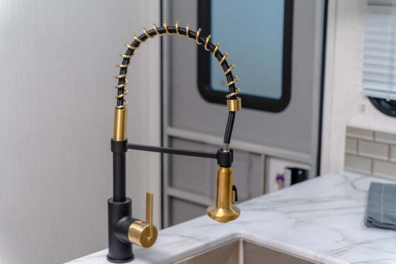 A modern kitchen faucet with a black and gold spring design and a pull-down spray head is installed over a marble countertop and stainless steel sink. A gray door and towel are in the background.