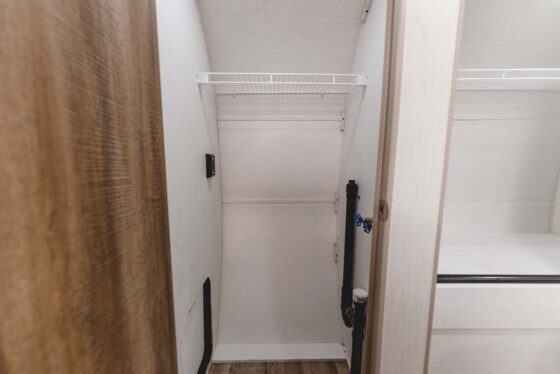 Empty white closet with a wire shelf and wooden floor, located next to a bed with a white mattress. A brown wooden door is partially visible on the left side of the image.