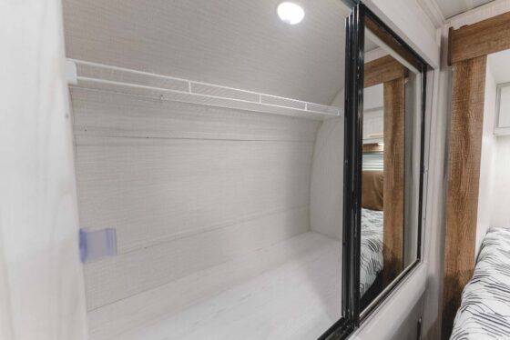 A view of an empty closet in an RV with a white wire shelf, light-colored walls, and a large mirror sliding door reflecting the adjacent bed and wood paneling.