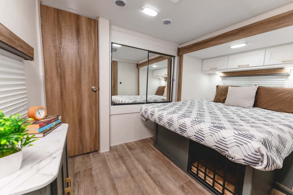 A cozy RV bedroom with a bed covered in geometric-patterned bedding, brown pillows, overhead cabinets, a large mirror, wood-style flooring, and a small table with a clock, books, and a potted plant.