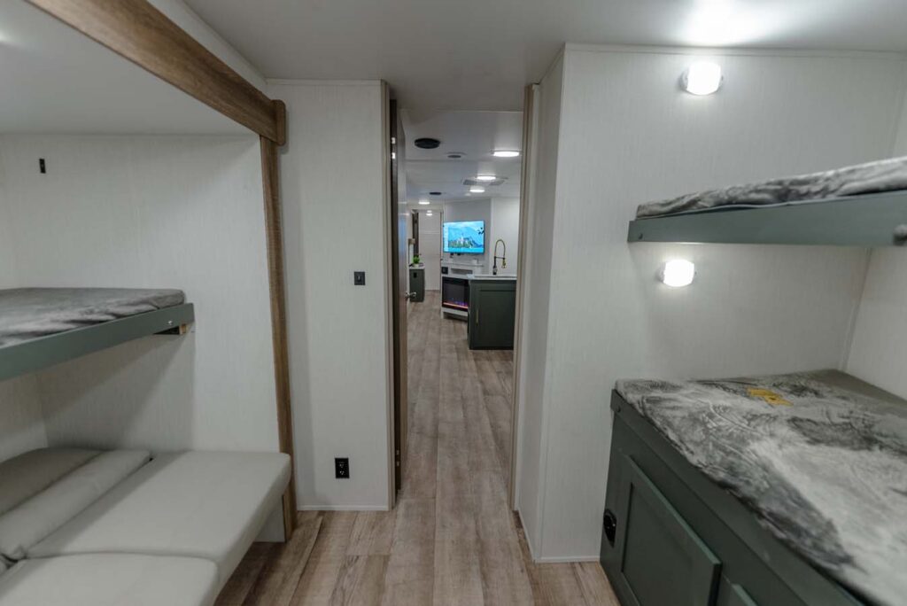 Interior view of a modern RV with bunk beds on the left and right, light wood floors, white walls, and a kitchen area with a TV visible in the background.