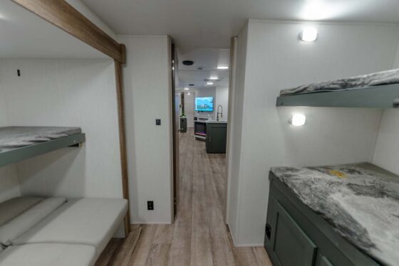 Interior of a modern RV showing bunk beds on both sides, a small couch, wood-look flooring, and a view into a kitchen area with a TV in the distance. The lighting is bright and the design is minimalist.