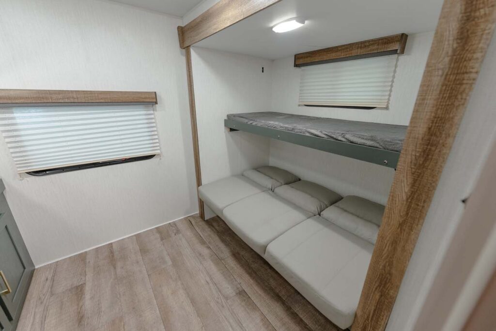 A compact room with light wood flooring, a window with blinds, and a bunk bed; the lower bunk is folded into a sofa, while the upper bunk has a thin mattress.