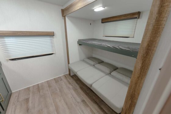 A small, modern RV bedroom with light wood flooring, a window with a closed blind, and a bunk bed featuring a single upper bunk and a lower bunk with three gray cushions.