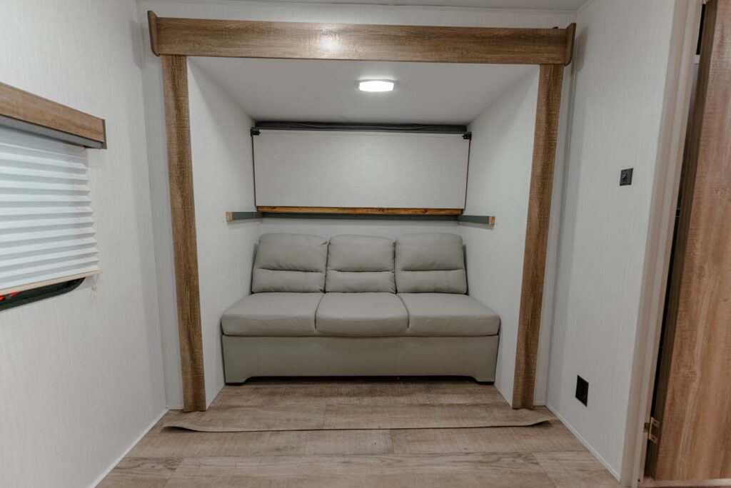 A small, modern room with wood accents featuring a light gray, three-seater sofa under a pull-down Murphy bed, set against a white wall and wood-look flooring.