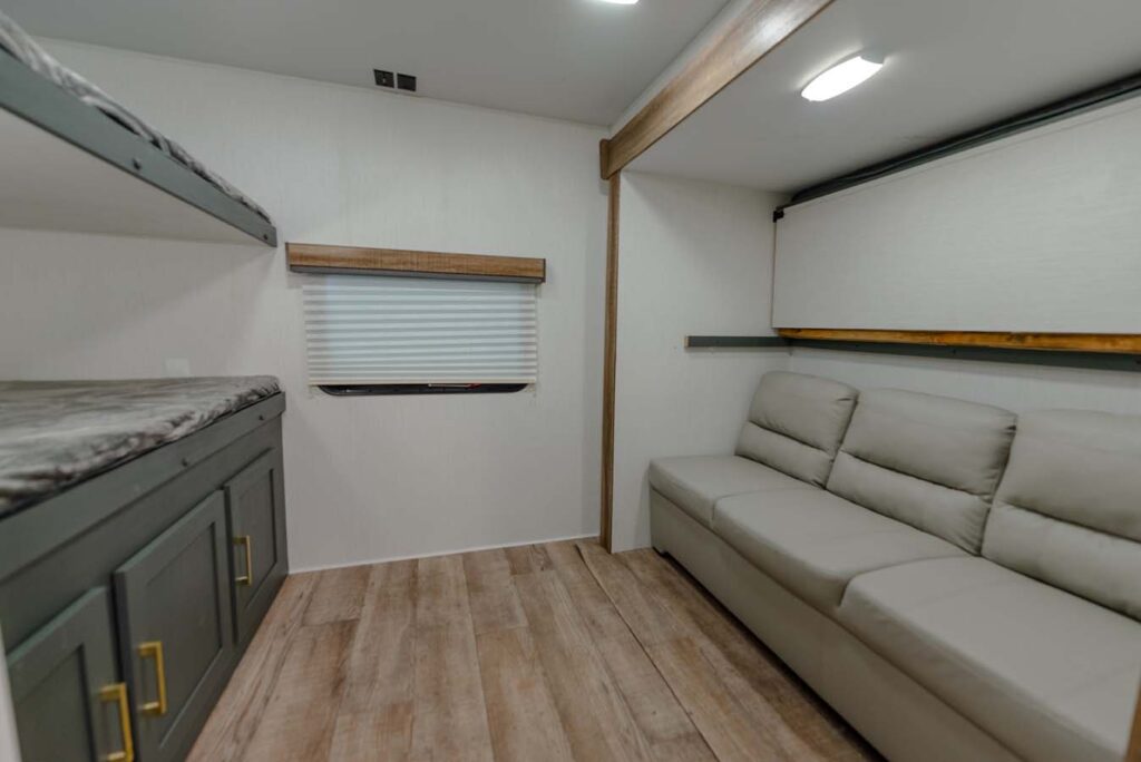 Modern RV interior with light wood flooring, a gray sofa on the right, and bunk beds with green cabinets below on the left. A window with a closed white blind is on the back wall. The space is tidy and minimal.