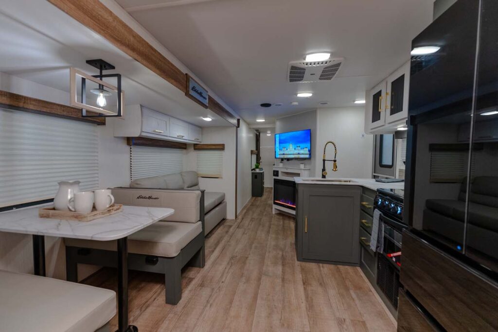 Modern RV interior with wood-style flooring, a dining area with a marble-like table, sofa, TV, kitchen with appliances, and contemporary lighting. The space is well-lit and features sleek, neutral decor.