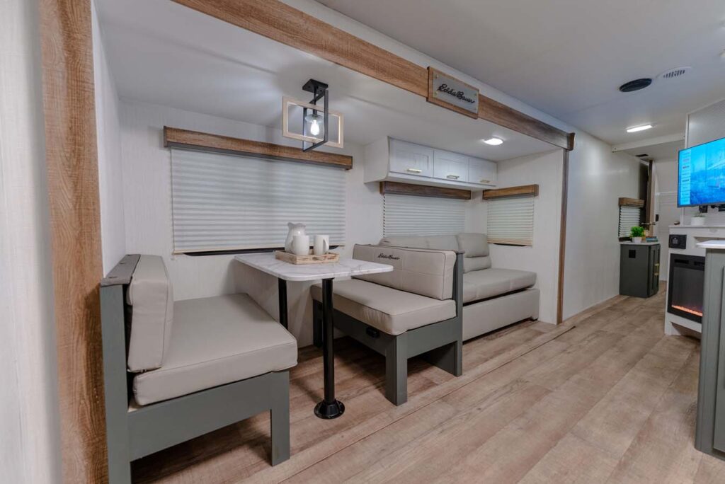 Modern RV interior with light wood floors, a dining area with gray cushioned benches and a white table set with a tray, teapot, and cups. Large windows have white blinds, and cabinets are above the seating.