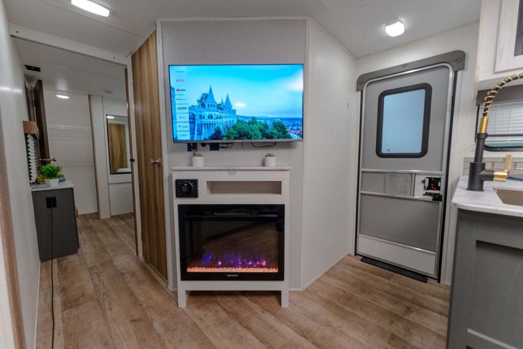 Modern RV interior featuring a mounted TV above an electric fireplace, wood-style flooring, a gray door, light-colored walls, and a kitchenette visible on the right. A hallway leads to additional rooms in the background.
