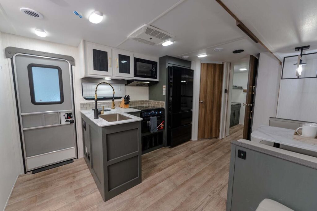 Interior of a modern RV with wood-style flooring, a kitchen featuring a sink, stove, microwave, and fridge, a dining booth to the right, and a door and hallway leading to other rooms in the background.